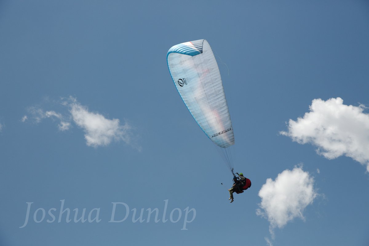 Image of a paraglider against a blue sky and white clouds with a grey watermark "Joshua Dunlop"